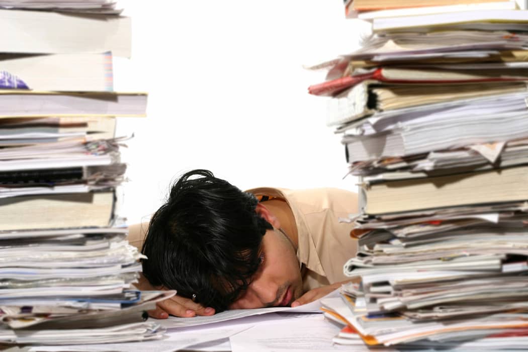 exhausted person with head down on desk next to two huge piles of folders and work