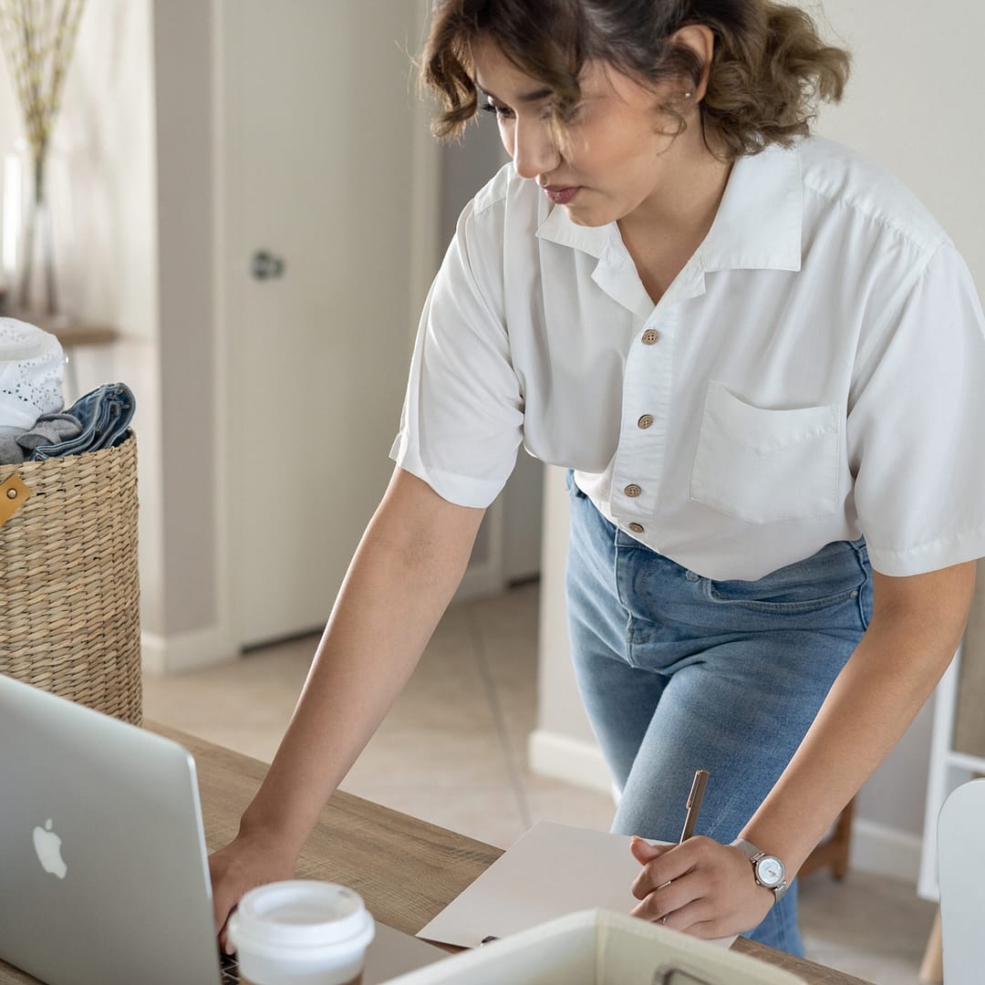 Woman in white shirt looking at a laptop while standing at a desk.